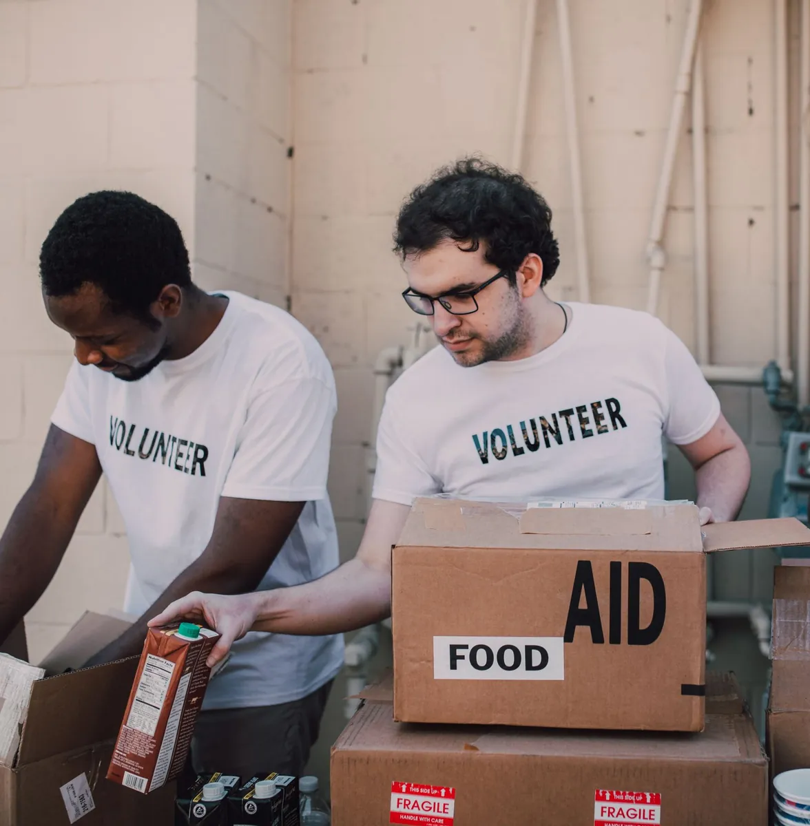 Two volunteers in white T-shirts packing a box labelled Aid and Food, one holding a carton of juice