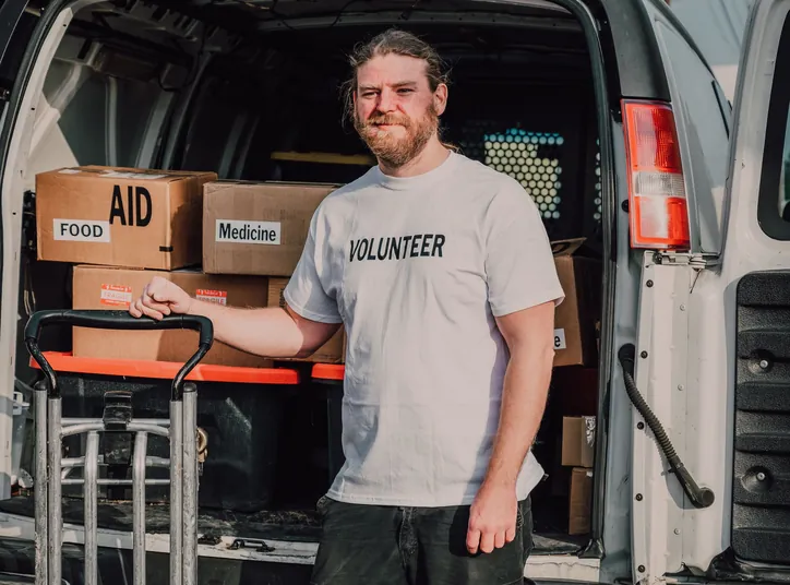 A bearded volunteer beside a hand truck at the open back of a van stacked with boxes marked Aid, Food and Medicine