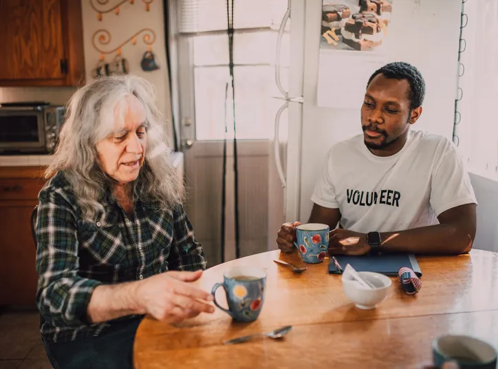 A volunteer and an older woman talking over mugs of coffee at a round kitchen table