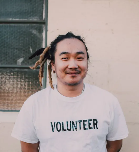 A smiling man with his dreadlocks tied back, in a white VOLUNTEER T-shirt, against a cream wall