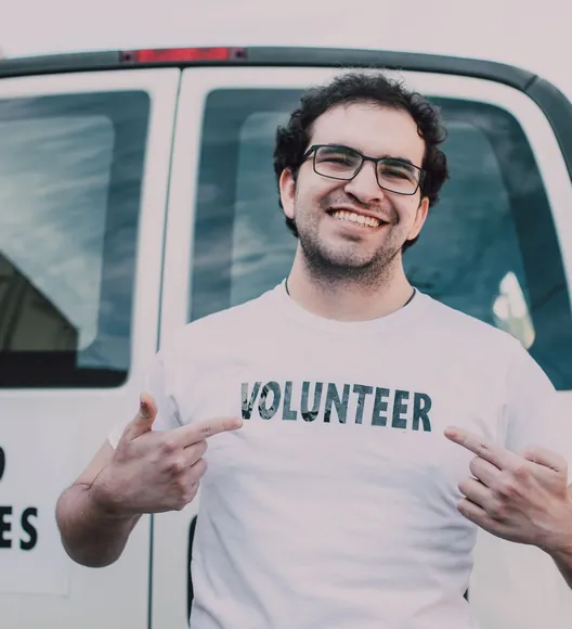 A man in glasses pointing at the word VOLUNTEER on his T-shirt, in front of a white van