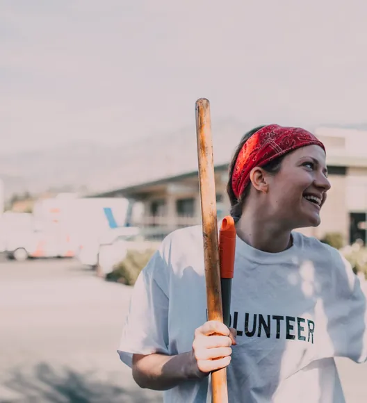 A woman in a red bandana and a white VOLUNTEER T-shirt laughing, holding a broom handle